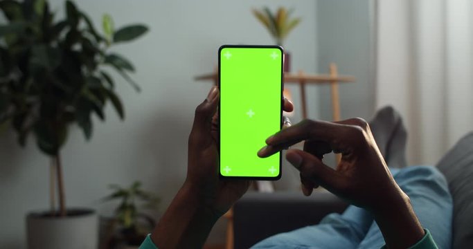 Afro american guy scrolling and touching mock up screen with trecking markers . Man using smartphone and browsing internet while lying on couch . Concept of chroma key and green screen.