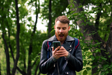 A guy with a thick beard opened his mouth wide in surprise while reading a message on a smartphone screen while standing in a dense forest.