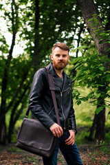 Fashionable guy with a red beard and a brown bag over his shoulder is standing in a green park on a background of trees.