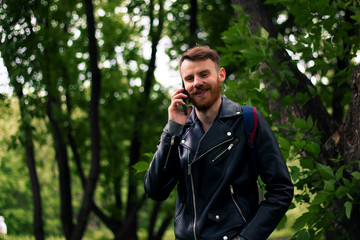 Smiling guy talking on a smartphone while standing in a forest in a leather jacket.