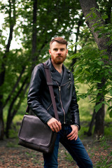 With his hand in his blue jeans pocket, a bearded guy in a black leather jacket is standing in the forest.