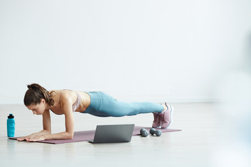 Naklejka premium Minimal full length portrait of fit young woman doing plank exercise while watching workout video via laptop at home, copy space
