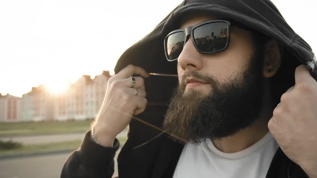 Portrait of bearded guy in black glasses and hood. Brutal stylish man with rings and signet on fingers is at sunset in city stands still in slow motion. Fashionable vogue person. Life style