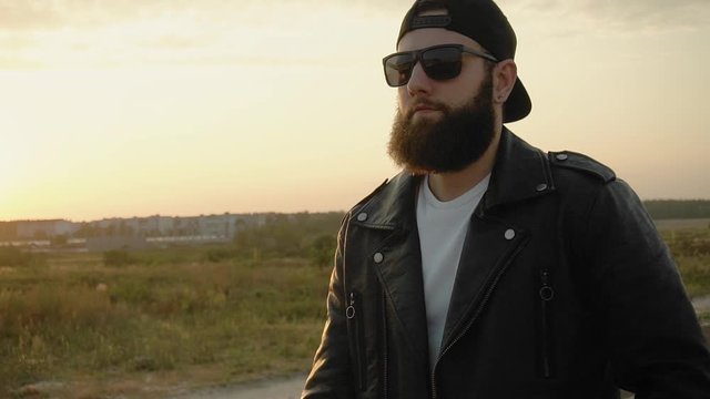 Bearded brutal image guy in black glasses, cap, leather jacket walks outdoors at sunset in slow motion. Fashionable stylish man goes in slow motion. Trendy person with earring. Vogue life style. Biker