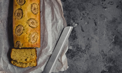 Homemade sliced healthy banana bread ready for serve on breakfast on the dark concrete background with craft baking paper and pastry knife. Top view. Free copy space for text