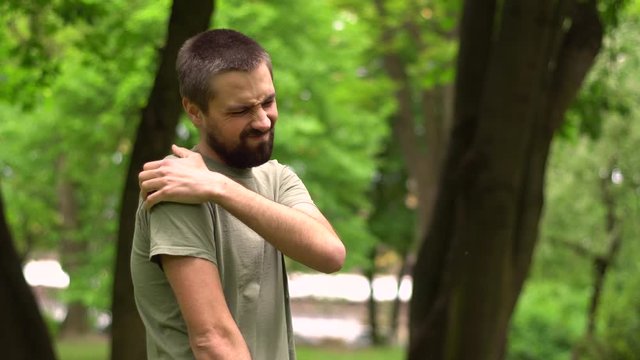 A young bearded man put his hand on his shoulder, which hurts him. Caucasian guy is standing in the park and on his face a grimace of pain.