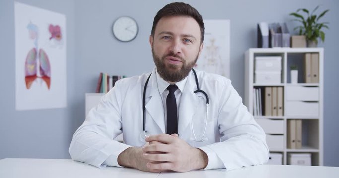 Medium plan of qualified experienced doctor in office in white doctor's gown with a statoscope speaking to client through a video conference. Web camera look. Сoncept of pandemic, modern medicine.