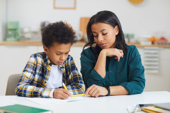 Portrait of cute African-American boy studying with female teacher watching him in class,