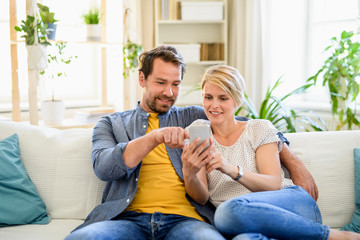 Happy couple in love sitting indoors at home, using smartphone.