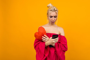sad caucasian blond girl in a red dress holding a phone and a red heart out of paper on an orange studio background