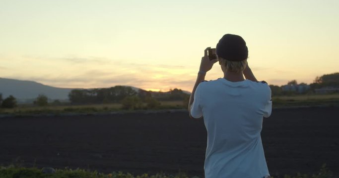 Back view of young man taking photos of sunset rural landscape with smartphone.