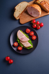 Sandwich with turkey ham meat, green salad and fresh cherry tomatoes slices on black plate near to ingredients on cutting board, blue minimal background, top view
