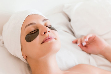 Fototapeta na wymiar Young woman with closed eyes and black eye patches is lying and relaxing in the bed after having a bath wrapped in towel. Beauty treatment and skincare concept