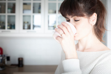 Pretty caucasian woman, with bangs and ponytail, drinking a cup of coffee in the morning in a white, bright lit, kitchen. Copy space. Relaxed and positive atmosphere.