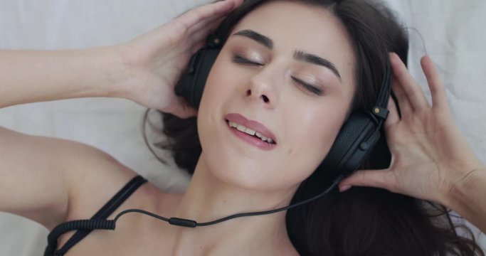 Top portrait of woman lying on bed, holds earcups, sways and bites lip at camera