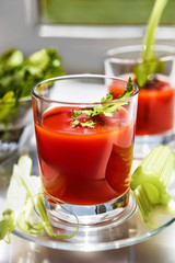 Two glasses of tomato juice with parsley and celery decorations, stand near the window, morning sunlight shines, shallow depth of field, selective focus. Natural drinks concept.