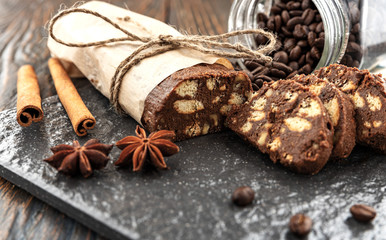 Chocolate salami on a stone slab, cinnamon and star anise sticks lie nearby, coffee beans pour out from a glass jar, shallow depth of field, selective focus. Breakfast dessert concept