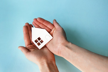 Hands holding white paper house in light blue background.