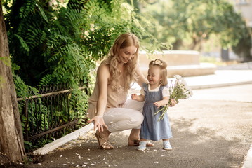 Young blonde mom and her one-year-old daughter sniff a bouquet of daisies in a summer park
