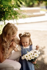Young blonde mom and her one-year-old daughter sniff a bouquet of daisies in a summer park
