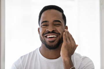 Headshot portrait of smiling young african American man apply nourishing facial cream or balm in...