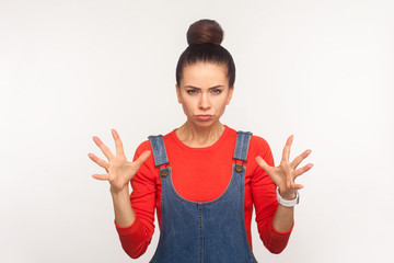 Portrait of irritated annoyed stressed girl with hair bun in denim overalls standing with clenched teeth and raised hands, expressing anger, aggression. indoor studio shot isolated on white background