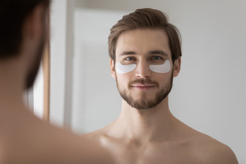 Portrait of young metrosexual man look in bathroom mirror apply use under eye anti-wrinkle patches, millennial male do morning facial procedures with hydrogel beauty product, skincare concept