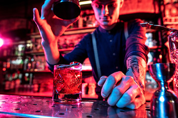 Close up of professional barman finishes preparation of alcoholic cocktail in multicolored neon light, gives it to client. Entertainment, drinks, service concept. Modern bar, trendy neoned colors.