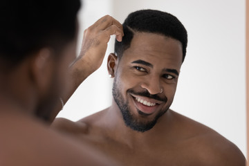 Smiling african American young man look in mirror in bathroom get ready in morning, happy biracial millennial male style hair, do daily beauty procedures treatment in bath, haircare concept