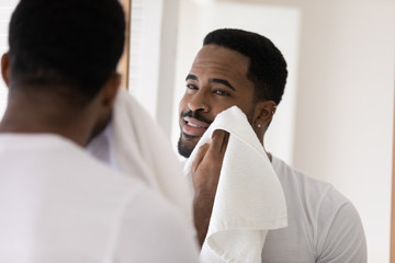 Handsome young african American man look in mirror in bathroom wipe rub face with towel after shower, millennial biracial male do morning beauty procedures, wash in bath, hygiene, skincare concept