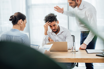 Selective focus of businessman quarreling at sad african american colleague near laptop and...