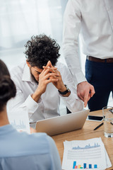 Selective focus of businessman pointing at laptop to sad african american colleague in office