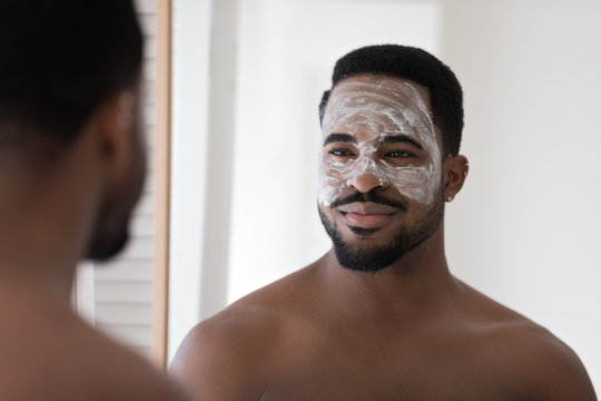 Handsome young african American man look in mirror in bathroom use facial blackhead pore removal mask, millennial metrosexual biracial male do morning beauty procedures in bath, skincare concept