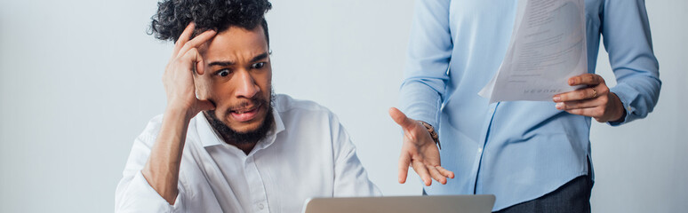 Panoramic shot of scared african american man looking at laptop near businesswoman holding documents