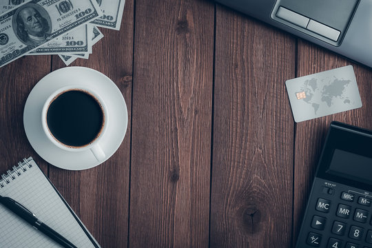Top view on a wooden rustic table with a cup of coffee, a credit card, a calculator, a notebook with a pen, dollar bills and a laptop with copy space.