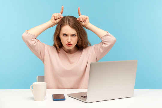 I'll Attack You! Aggressive Young Woman Employee Sitting At Workplace With Laptop And Doing Bull Horn Gesture, Threatening, Looking Angrily At Camera. Indoor Studio Shot Isolated On Blue Background