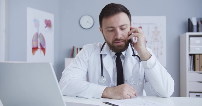 Medium plan of young beautiful male doctor in lab coat and statoscope in front of laptop talking on mobile phone while working at desk in medical office. Сoncept of pandemic, modern medicine.