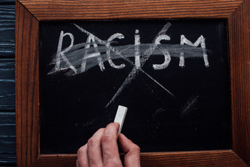 Top view of man holding chalk near blackboard with racism lettering on dark wooden background