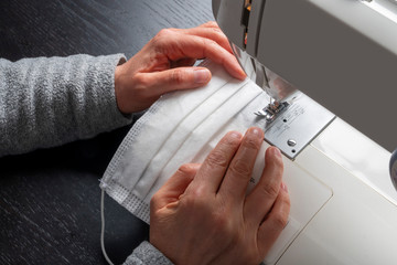 Obraz na płótnie Canvas Corona virus face protection mask sewing concept: view from behind on hands holding vertical a white cotton material on sewing machine for a DIY medical mask activity at home