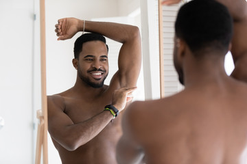 Smiling young naked african American man look in mirror in bathroom after shower apply deodorant on...