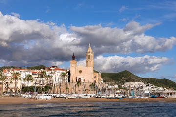 Beautiful view of the church Sant Bartomeu and Santa Tecla in Sitges with boats on the beach under the beautiful sky.