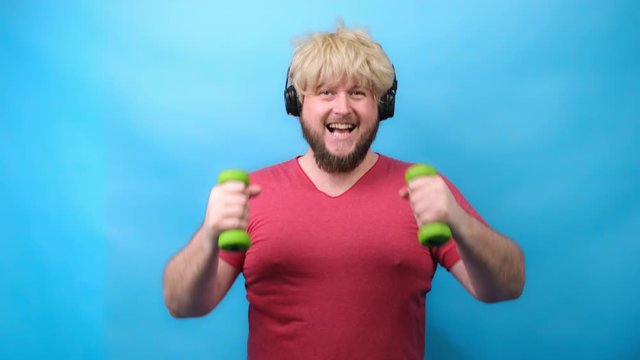 Funny man freaks in headphones and with a haircut from 70 sin pink T-shirt deals with small green dumbbells and smiles