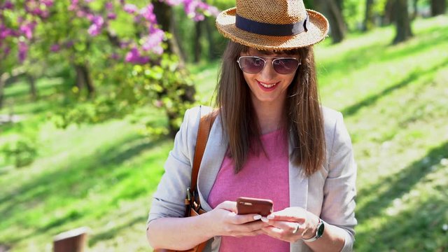 Young woman in hat using phone outdoor in park pink blossom on tree.