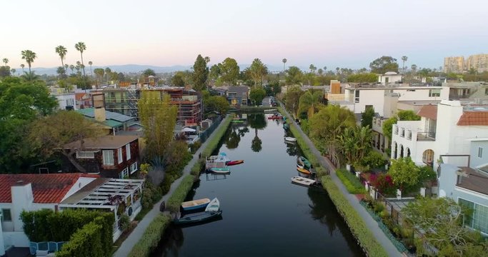 Aerial, rising, drone shot tracking forward and tilting down towards canoes and boats on a still Venice Canal, in the middle of colorful houses, empty for CoronaVirus, in Los Angeles, California, USA