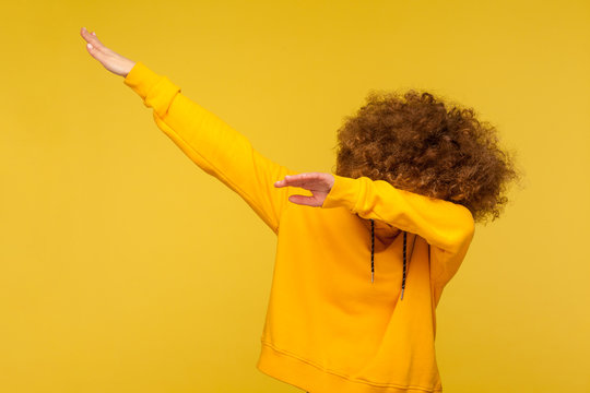 Portrait of curly-haired young woman in urban style hoodie hiding face with dab dance move, performing internet meme of success, dabbing trends. indoor studio shot isolated on yellow background