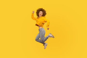 Fototapeta na wymiar Portrait of enthusiastic joyful curly-haired girl in urban style hoodie and jeans jumping high in air, flying and shouting with amazed happy expression, celebrating success. studio shot isolated