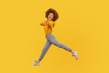 Fototapeta na wymiar Hey you! Portrait of lively energetic curly-haired girl in urban style outfit walking through air in wide strides, pointing to camera and shouting message. studio shot isolated on yellow background