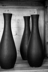 Three empty black ceramic vases on wooden shelf against concrete wall. Black and white image.