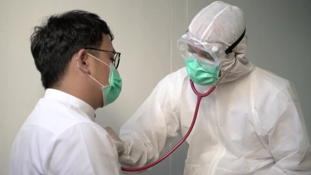 Coronavirus concept. Asian male doctor in a protective suit, protective mask and medical glasses listens to the breath of an elderly sick man using a phonendoscope. 4K Resolution.