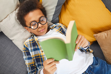 Above view portrait of cute African boy wearing big glasses reading book while lying on couch,...